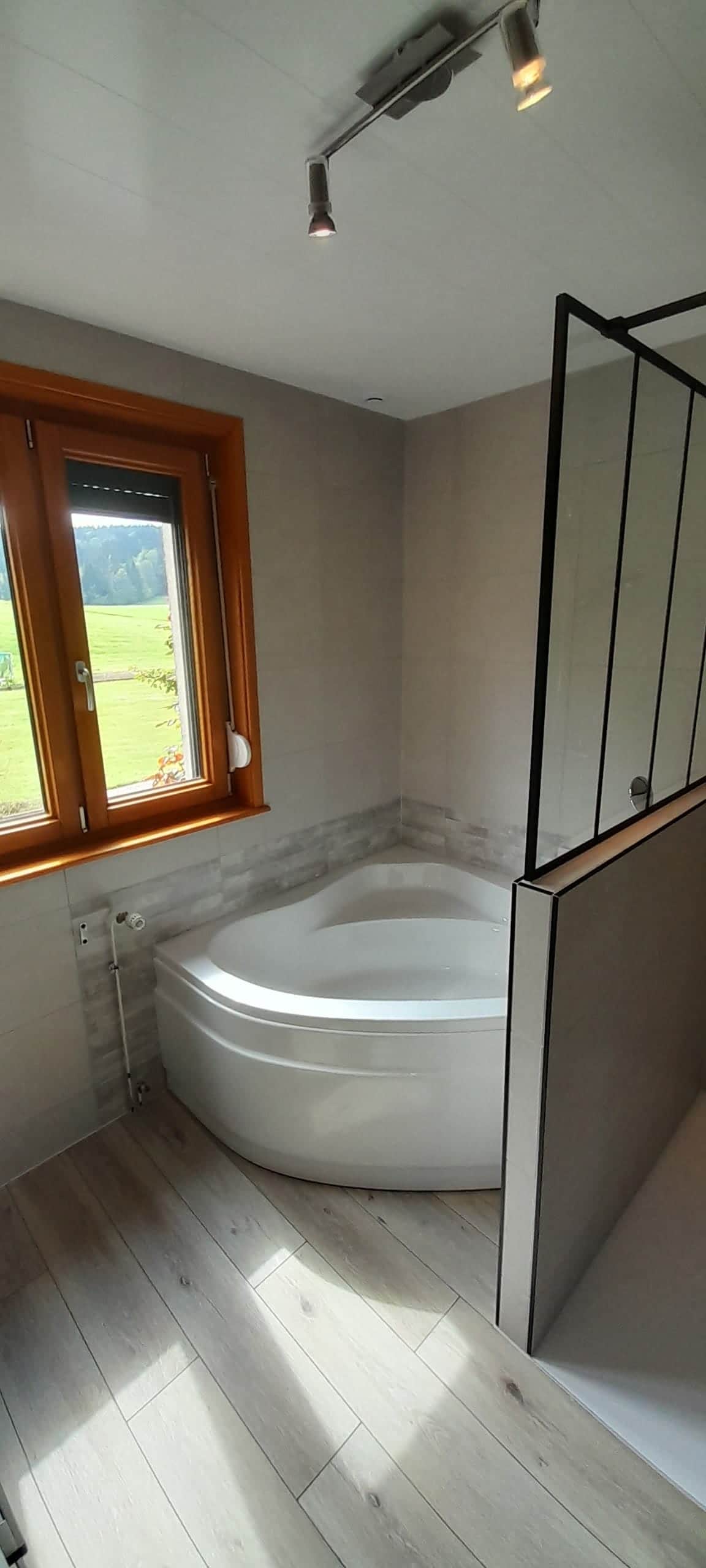 Salle de bain rénovée avec baignoire d'angle, paroi verrière noire et sol imitation parquet clair