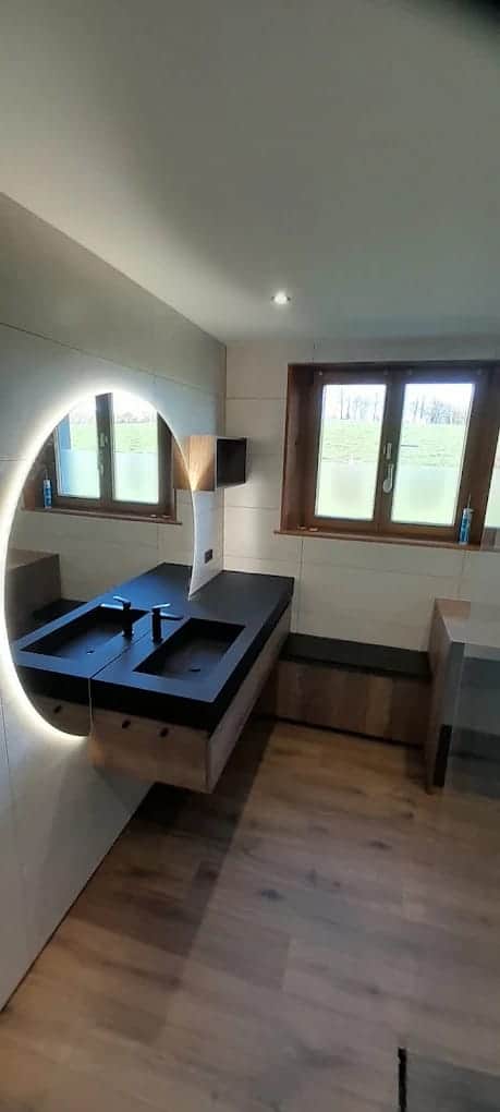 Salle de bain avec plan double vasque noir suspendu et miroir rond rétroéclairé, réalisée par AML Plomberie